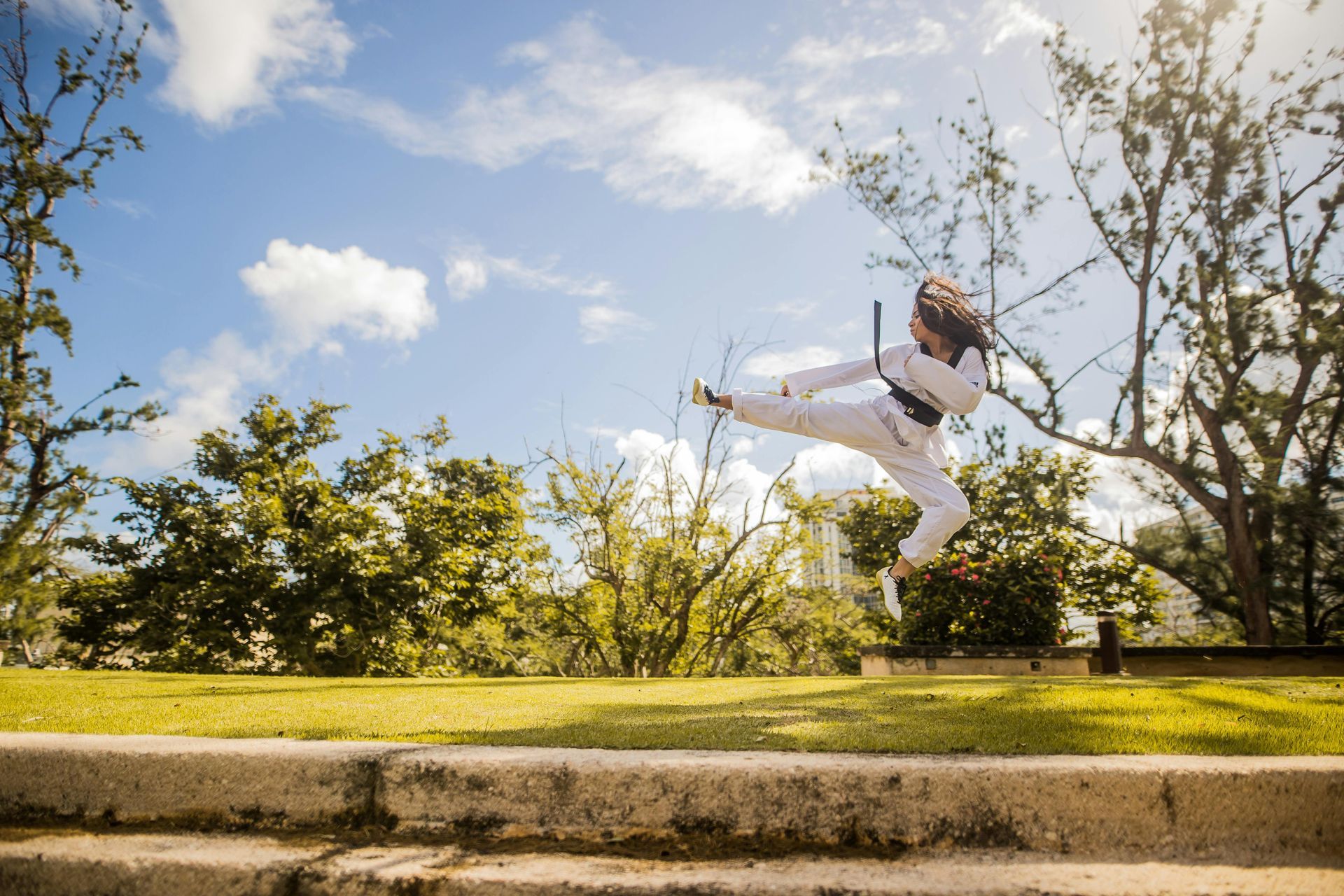 Person in white martial arts uniform performs a high kick outdoors on a sunny day.