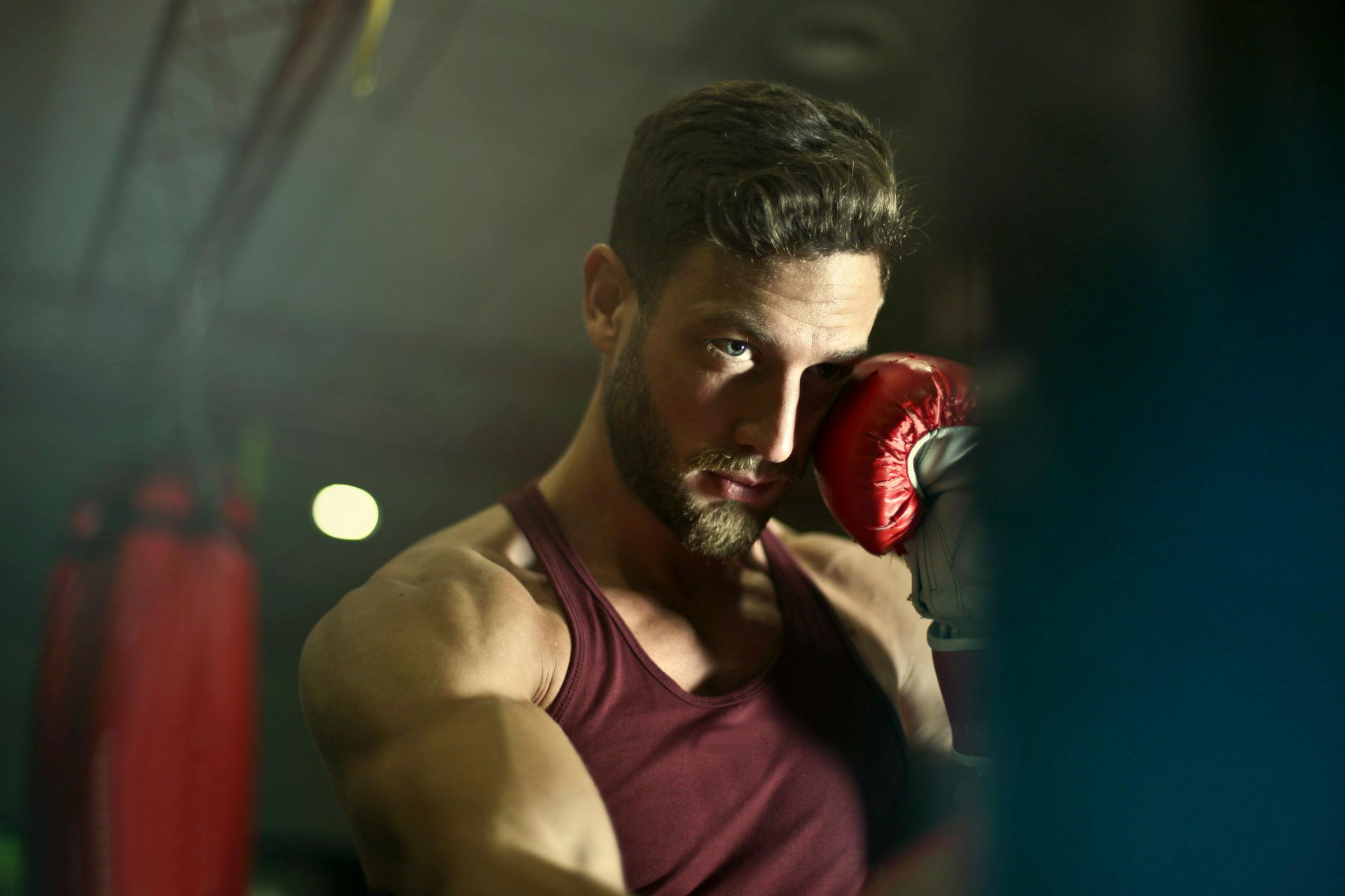 Boxer in a maroon tank top throws a punch, wearing red gloves, in a dimly lit gym.