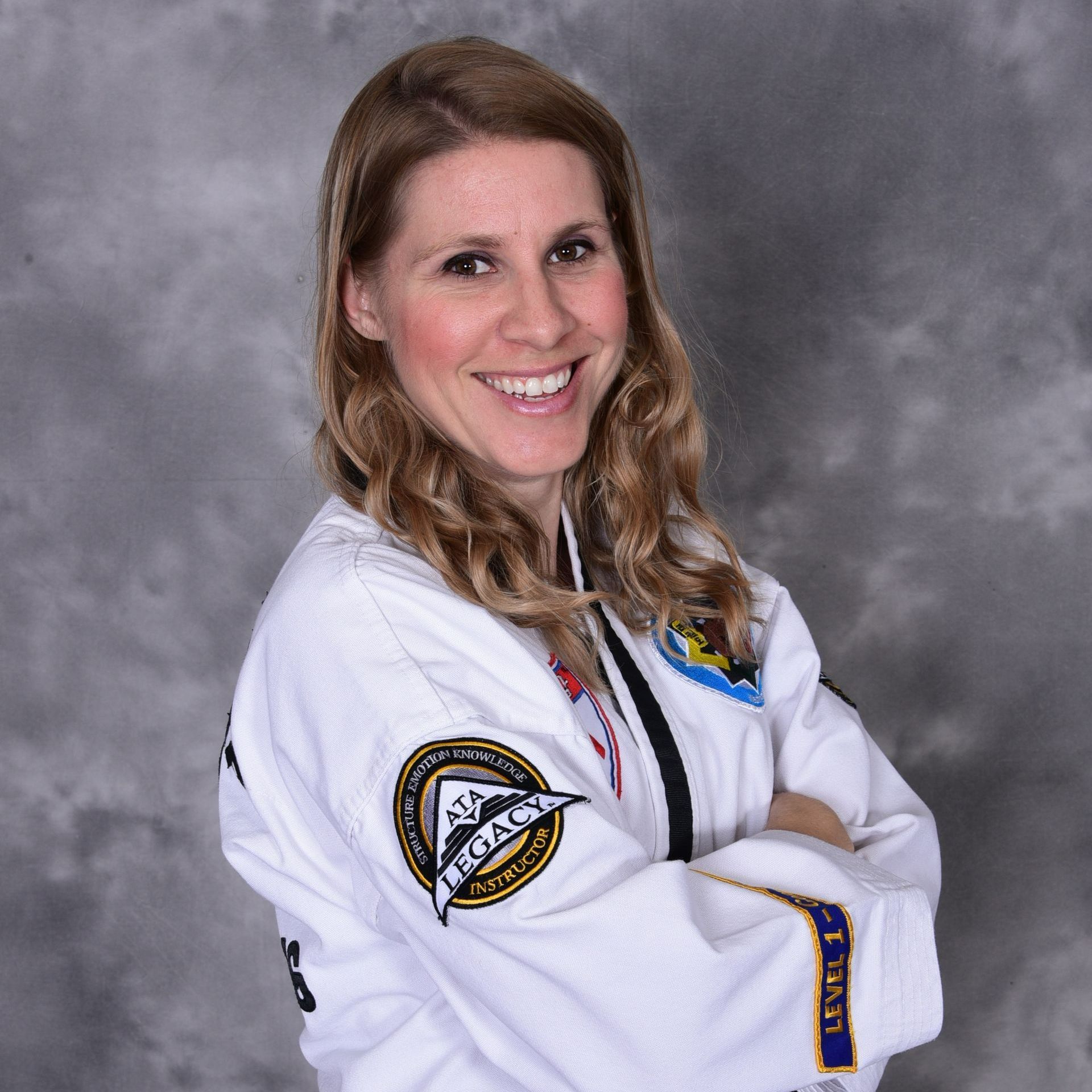 Woman in white martial arts uniform smiles, arms crossed. Background: gray mottled.