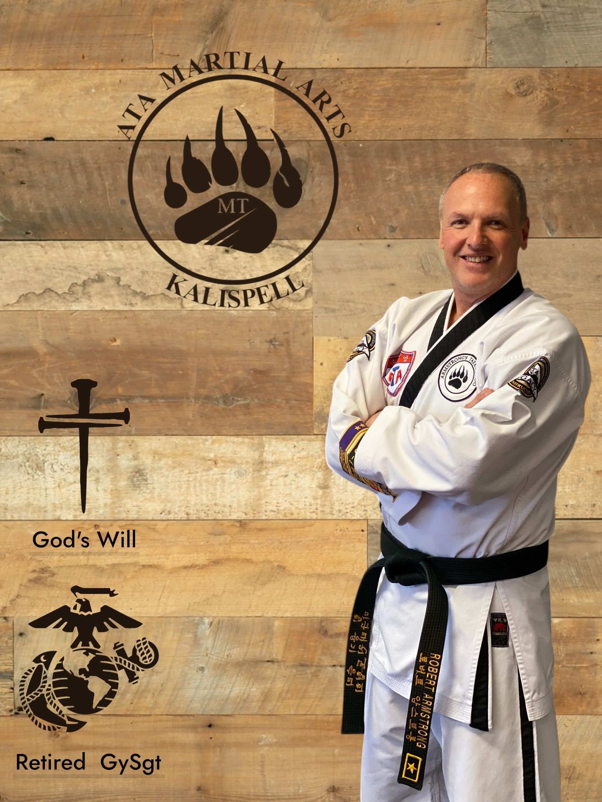 Man in martial arts uniform smiles, arms crossed, beside a wooden wall with logos.
