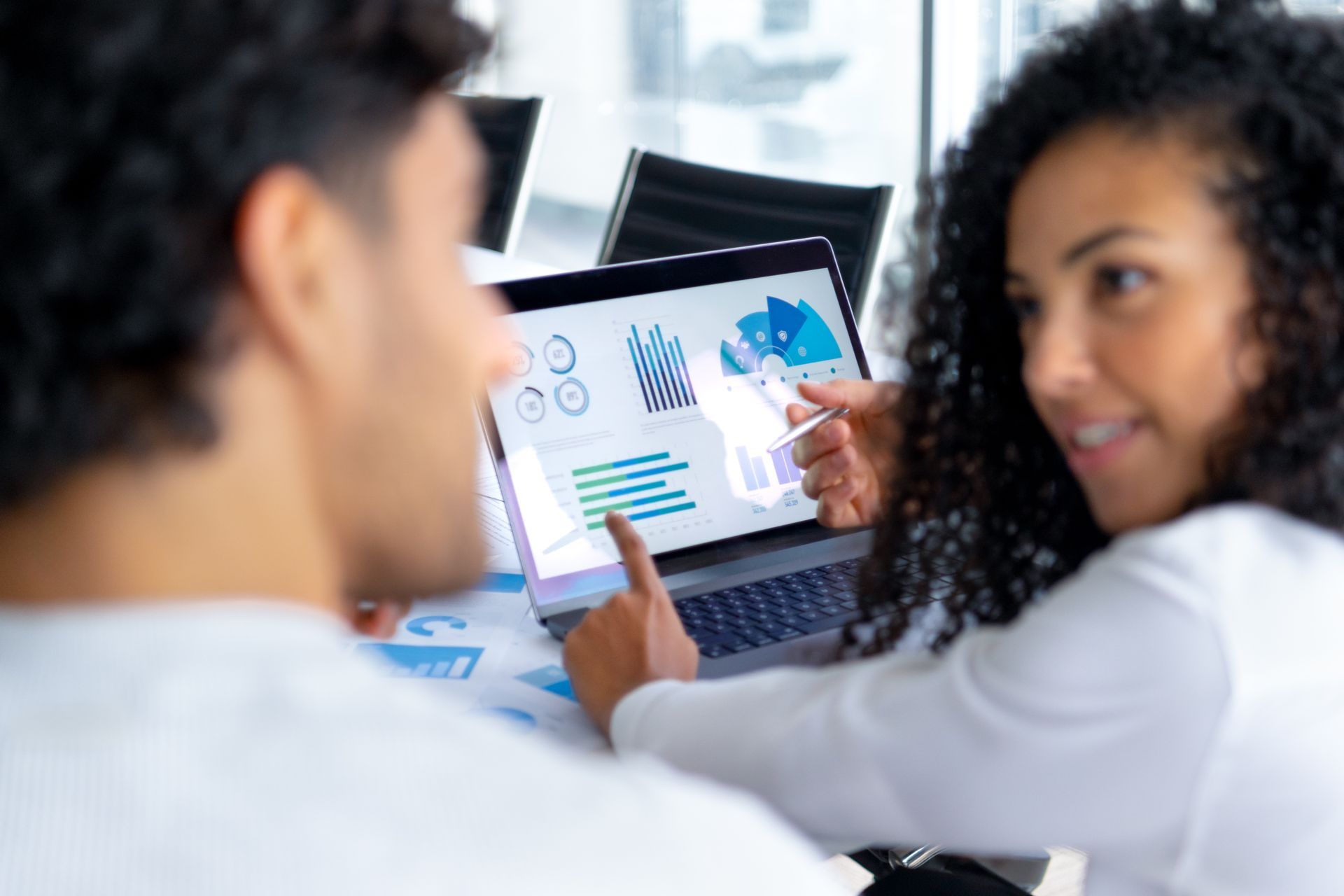 Two colleagues, smiling, discussing charts displayed on a laptop screen in a modern office.