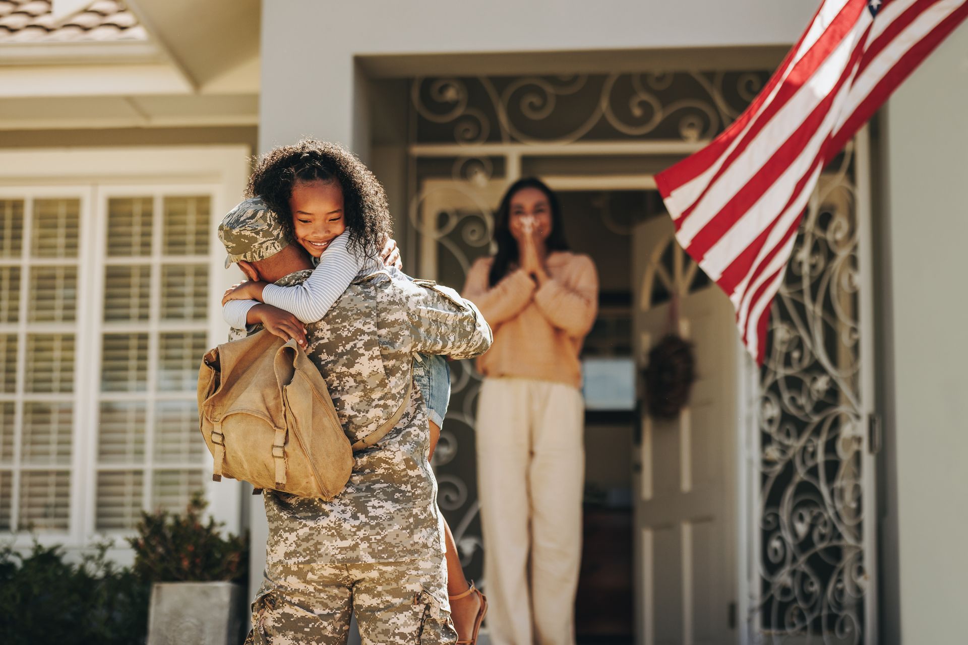 Soldier in uniform hugging child, with mother in the doorway, American flag, sunny day.