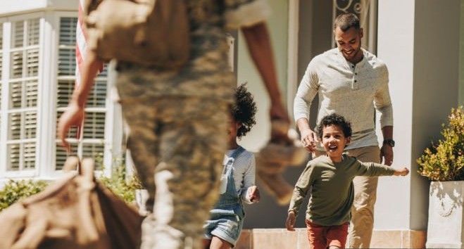 Soldier returns home, greeted by family outside a house; children excitedly run towards him.