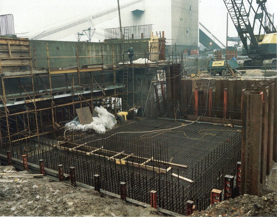 Construction site with steel rebar foundation, wooden formwork, cranes, and a steam plume in the background
