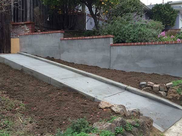 Concrete wheelchair ramp along a retaining wall in a yard, bordered by dirt and rocks