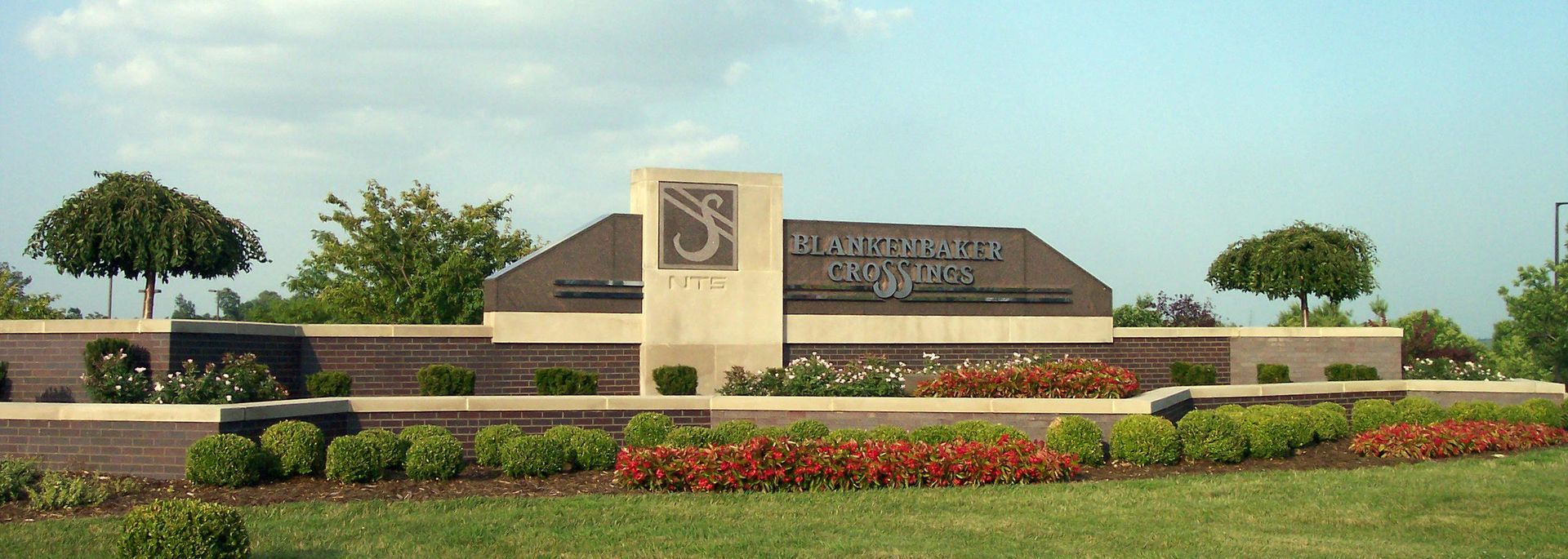 Landscaped campus entrance with a stone sign, flower beds, and trees under a clear blue sky