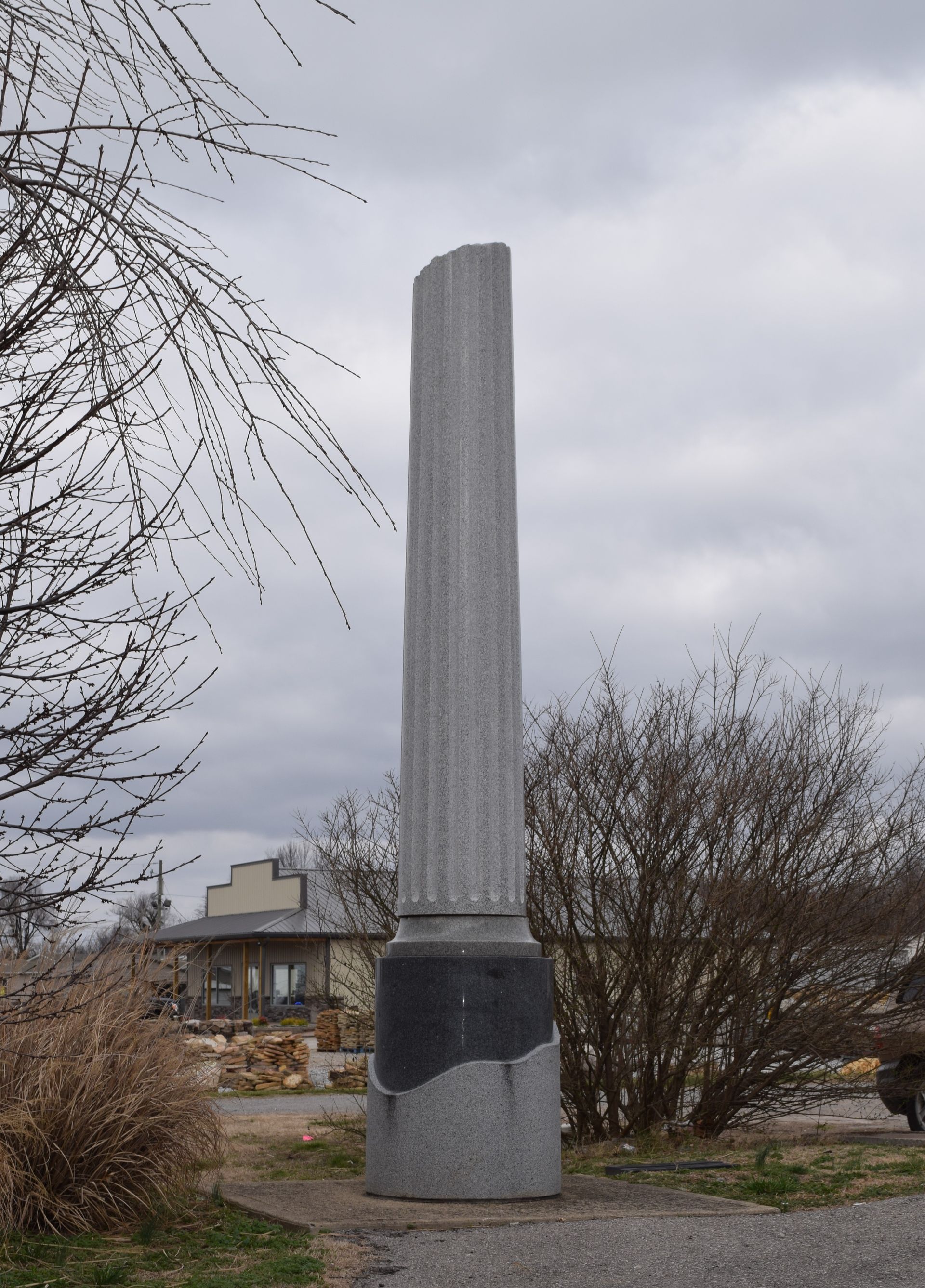 Tall gray stone monument in a cloudy outdoor park setting, with leafless trees and buildings in the background