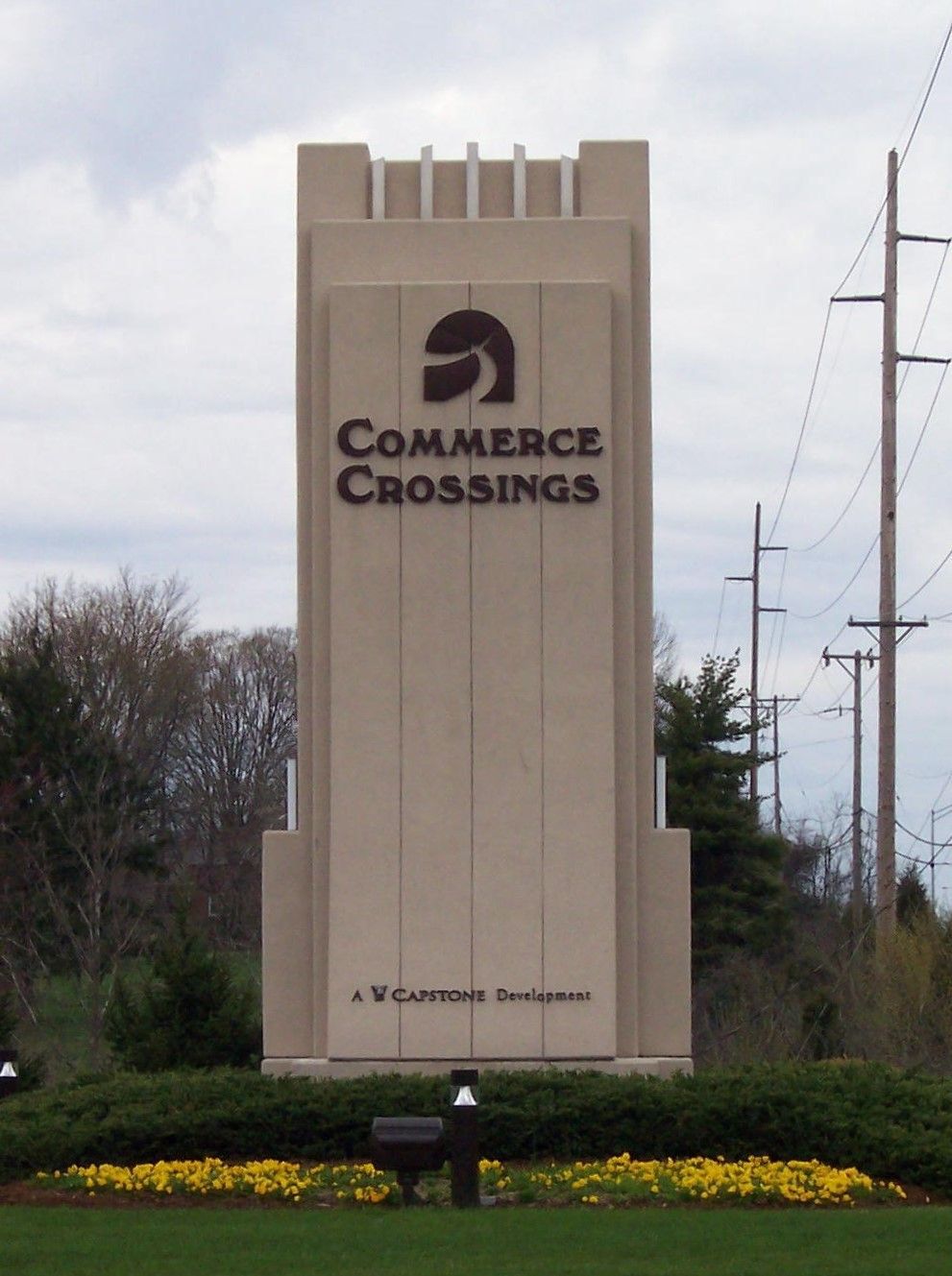 Tall beige Commerce Crossings monument sign beside yellow flowers and utility poles outdoors