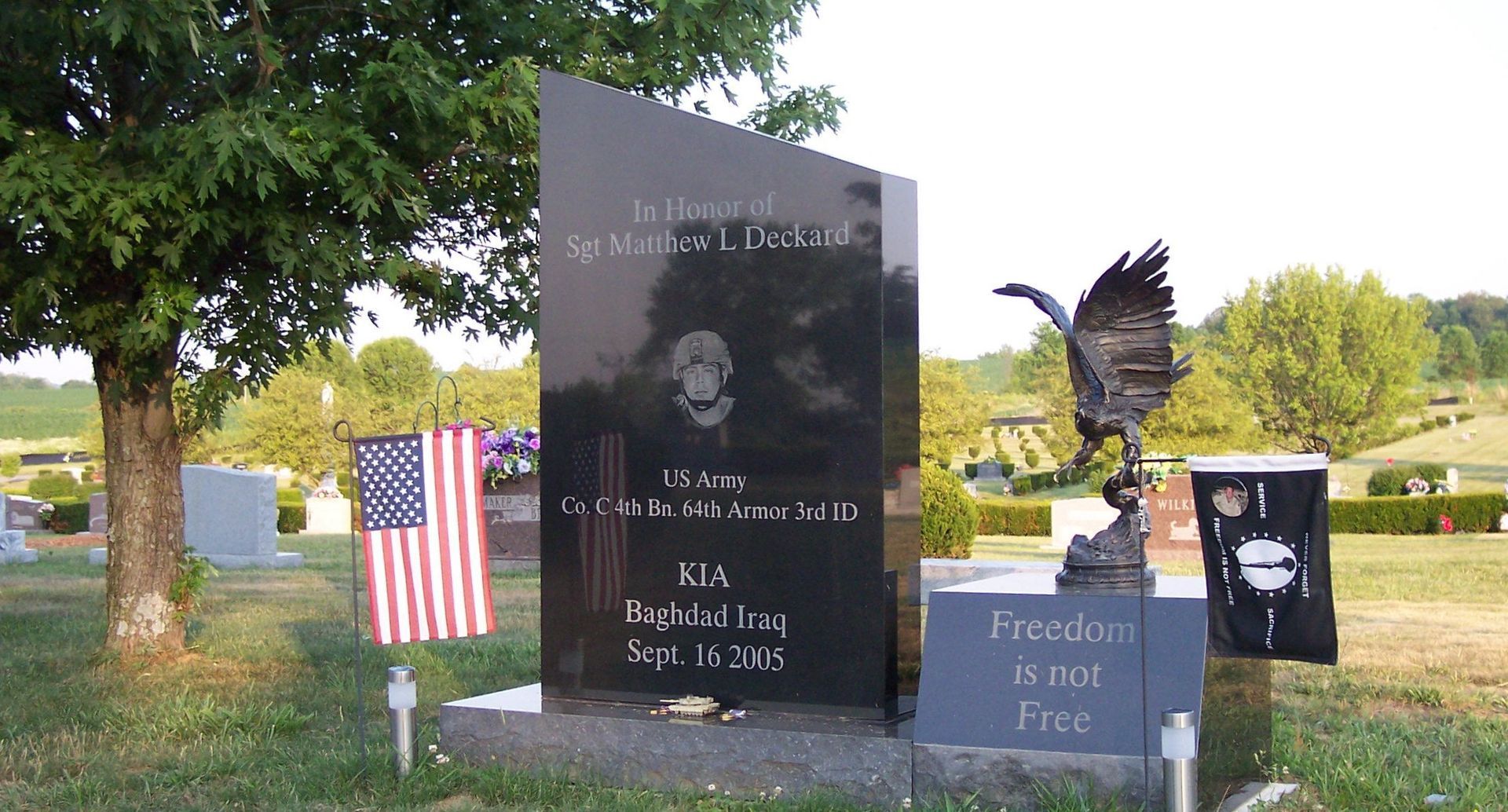 Memorial headstone in a cemetery with American flag, flowers, and an eagle statue beside it.
