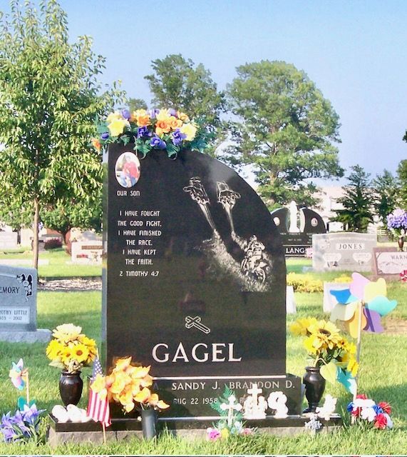 Black gravestone with flowers and pinwheels in a cemetery, inscribed Gagel and Sandy M. Alton.