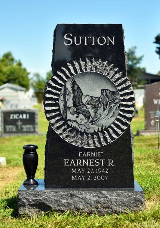 Black Sutton gravestone with engraved bird medallion and small vase in a cemetery