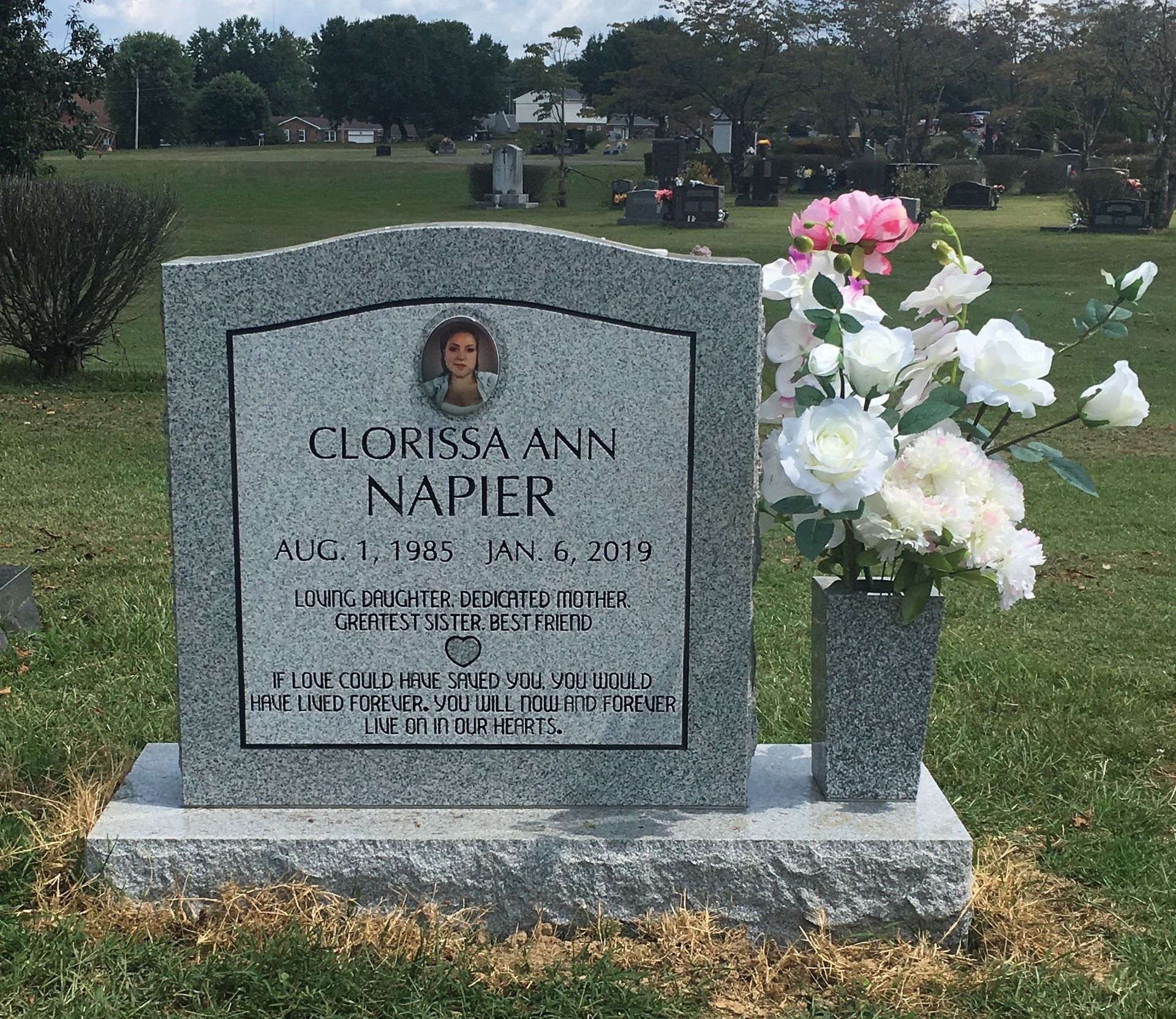 Gray gravestone with portrait plaque and pink-white flowers in a cemetery