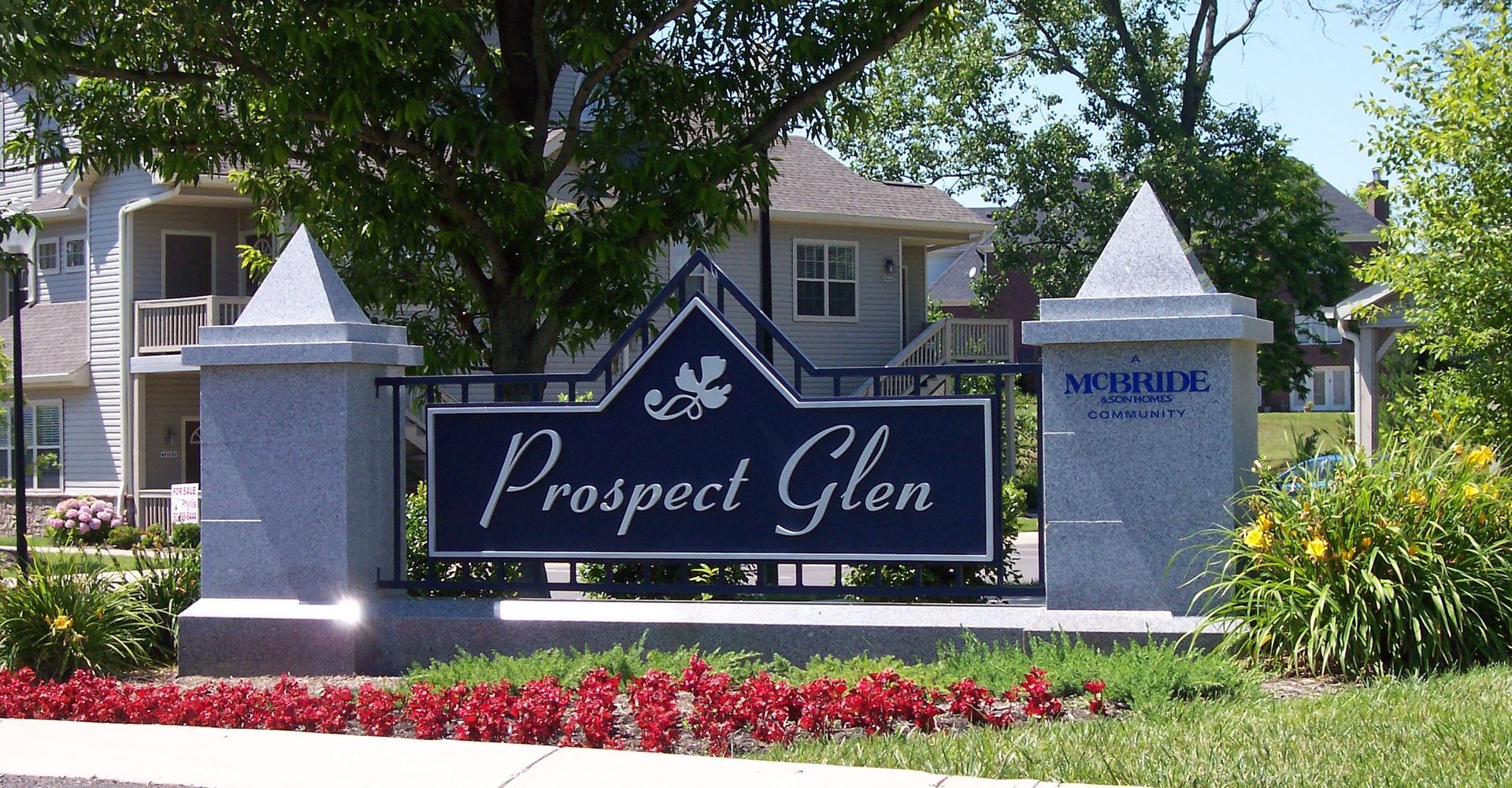 Entrance sign for Prospect Glen neighborhood with stone pillars, flowers, and trees in front of houses