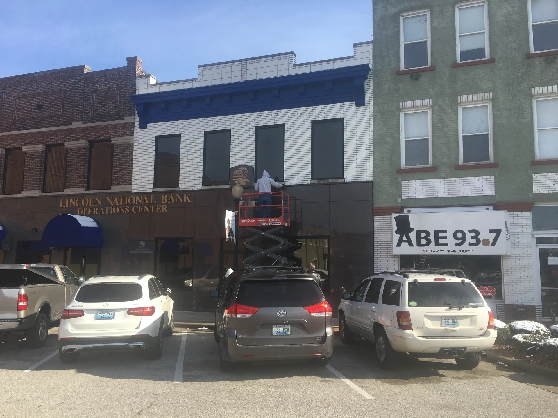 Street storefronts with parked cars and a sign reading “ABE 93.7”