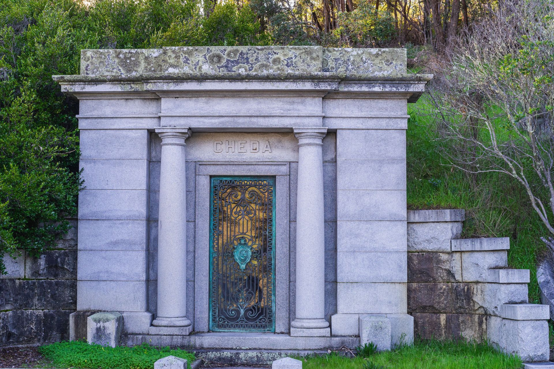 Stone mausoleum entrance with ornate bronze door and columns set against a wooded hillside