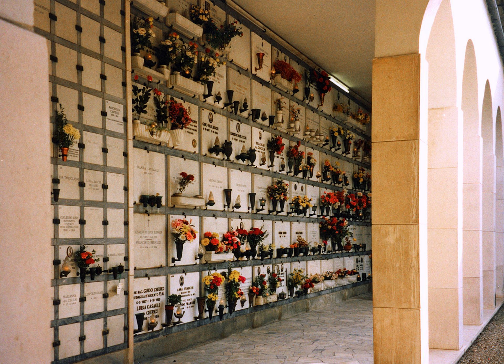 Wall of cemetery niches with flowers and memorial plaques under an arched colonnade