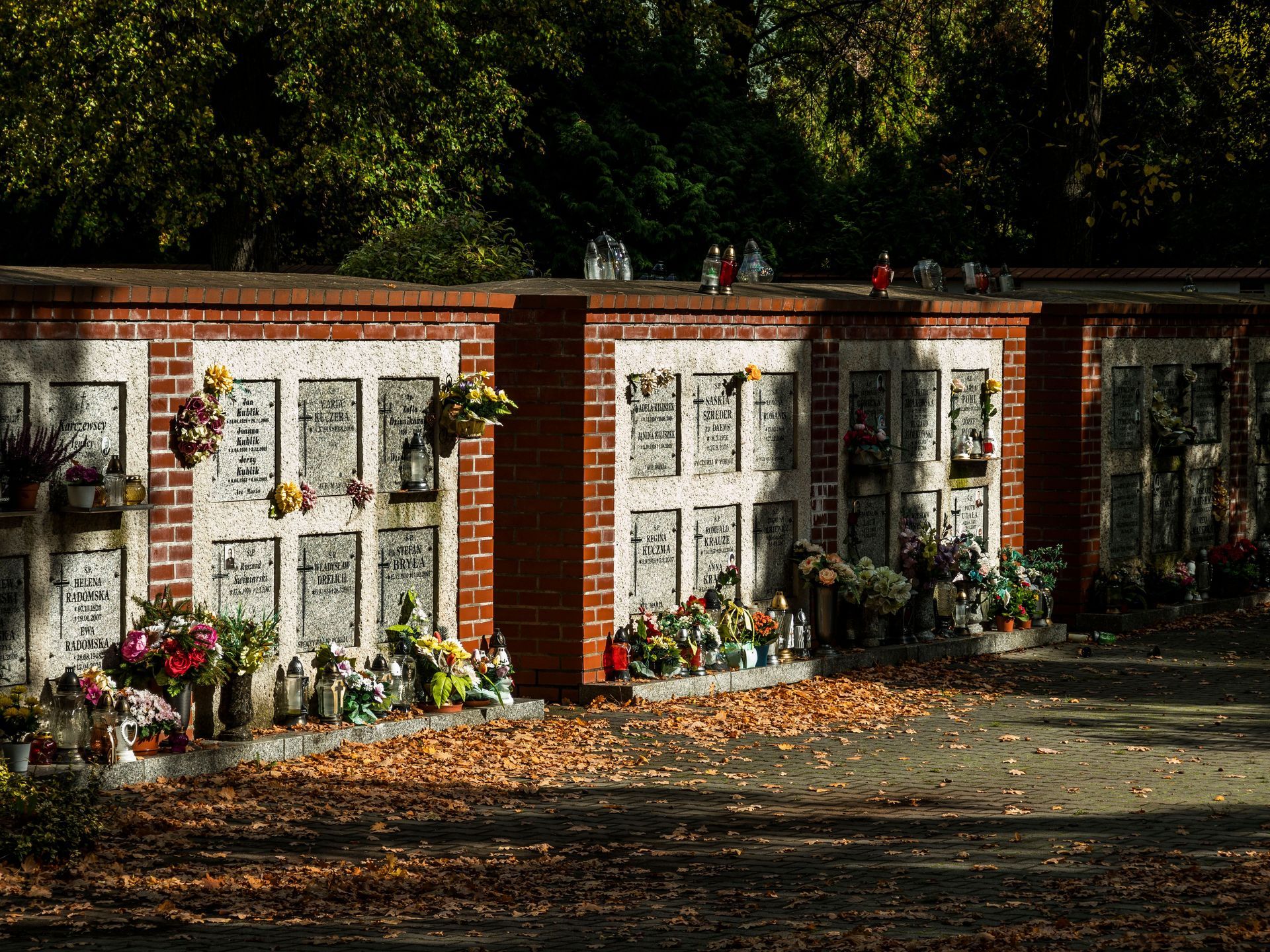 Brick memorial wall with white plaques, flowers, and a large tree overhead in a quiet outdoor setting
