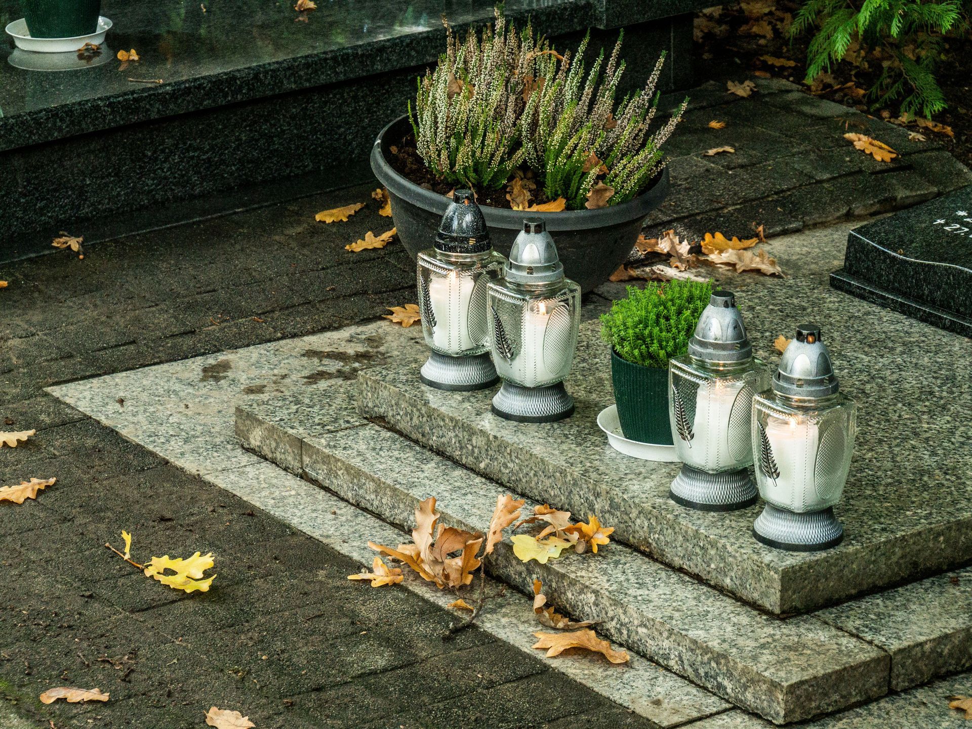 Small memorial with four silver lanterns and a potted plant on a stone slab beside fallen leaves.