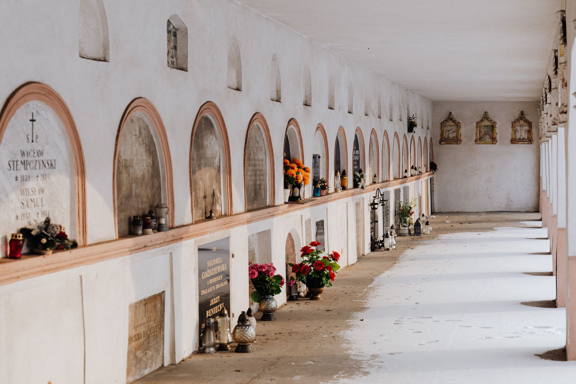 Long cemetery columbarium corridor with arched niches, flowers, candles, and sunlit white walls