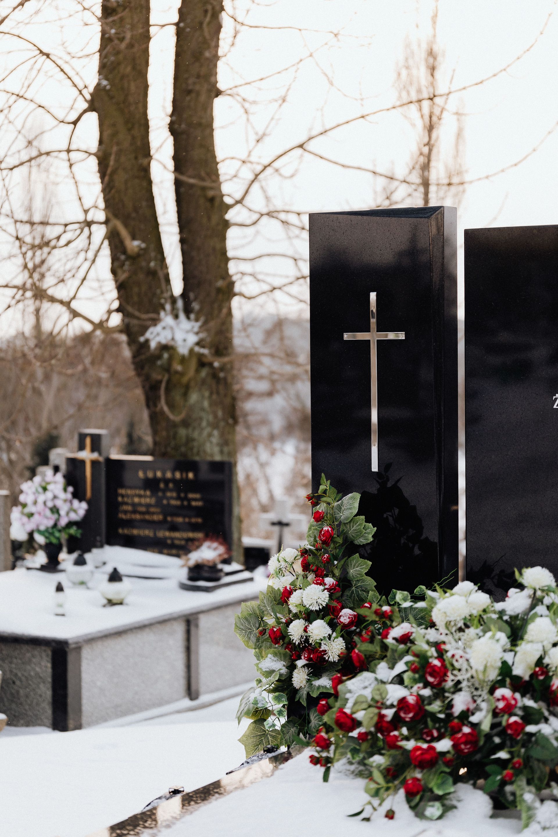 Snow-covered cemetery with black headstones, white crosses, and red-and-white flowers