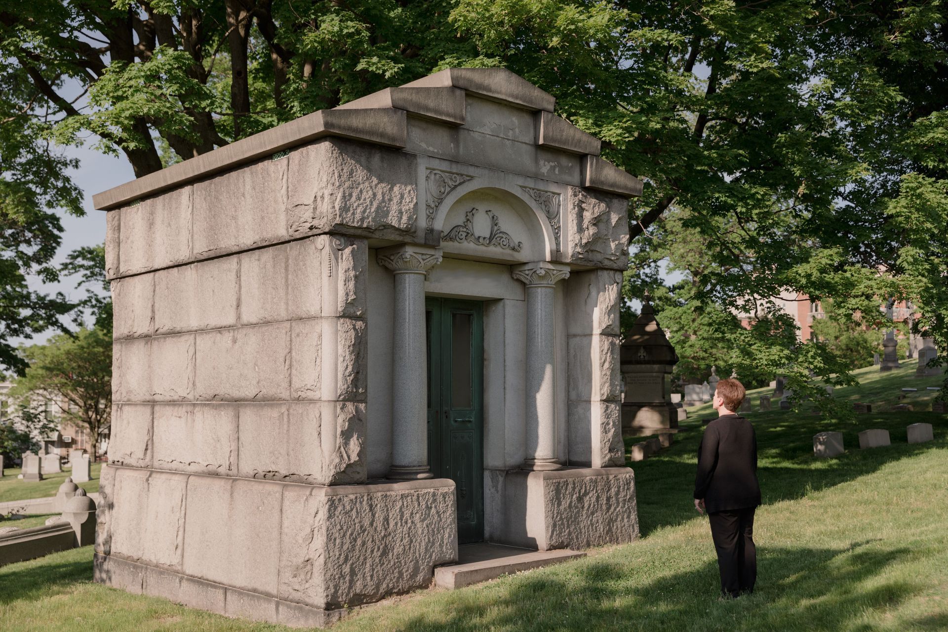 Stone mausoleum with a green door in a cemetery, with a person standing nearby under trees.