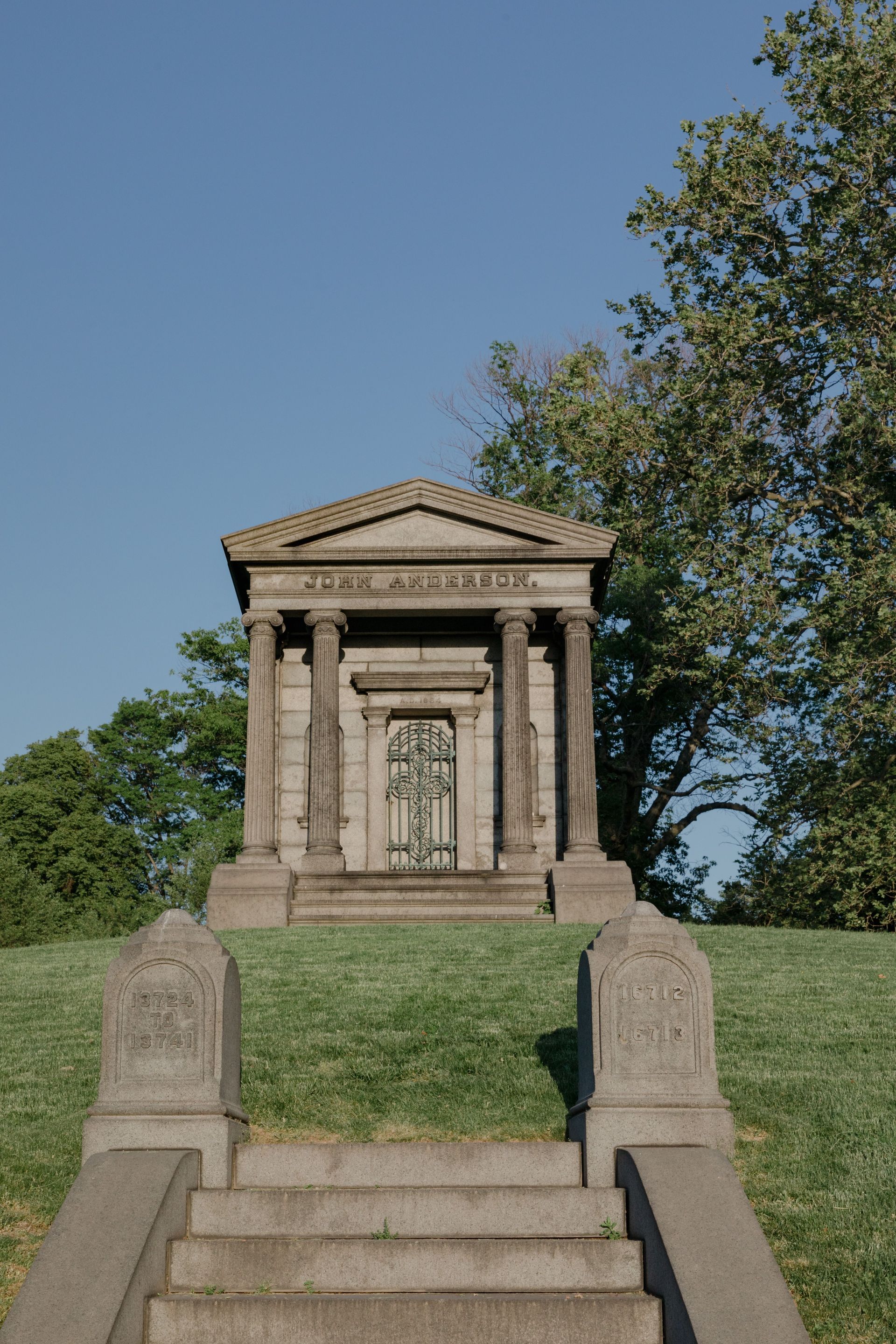 Stone mausoleum atop grassy hill, framed by steps and trees under a clear blue sky