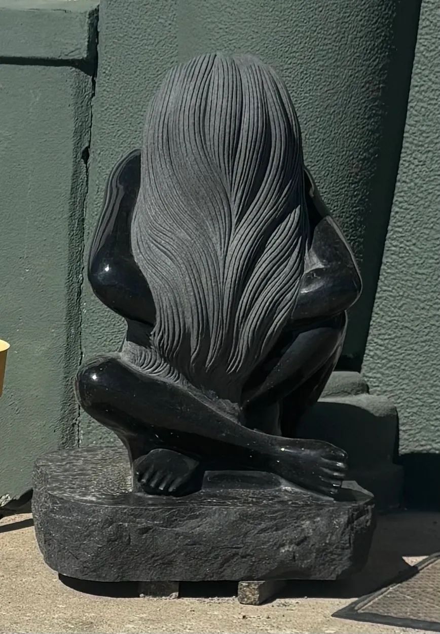 Black abstract sculpture on a stone base outdoors against a green wall.