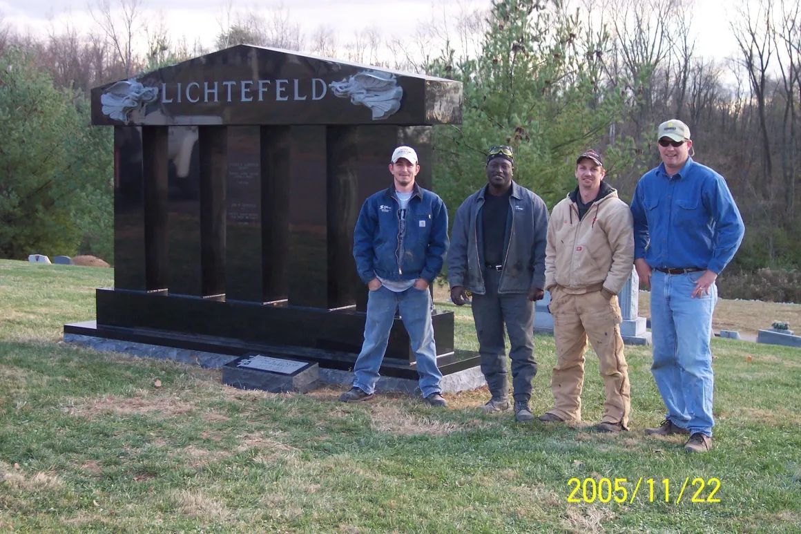 Four people standing outdoors beside a large dark monument with “Lichtfeld” on it.