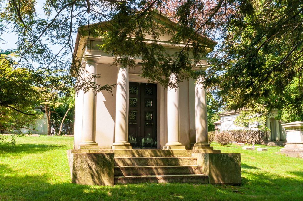 Small white columned pavilion in a sunlit green park, framed by trees and steps leading up to the entrance