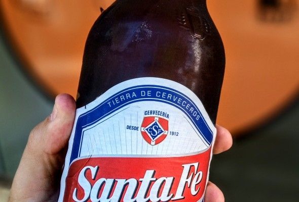 Mano sosteniendo una botella de cerveza Santa Fe; etiqueta roja, blanca y azul con logotipo.