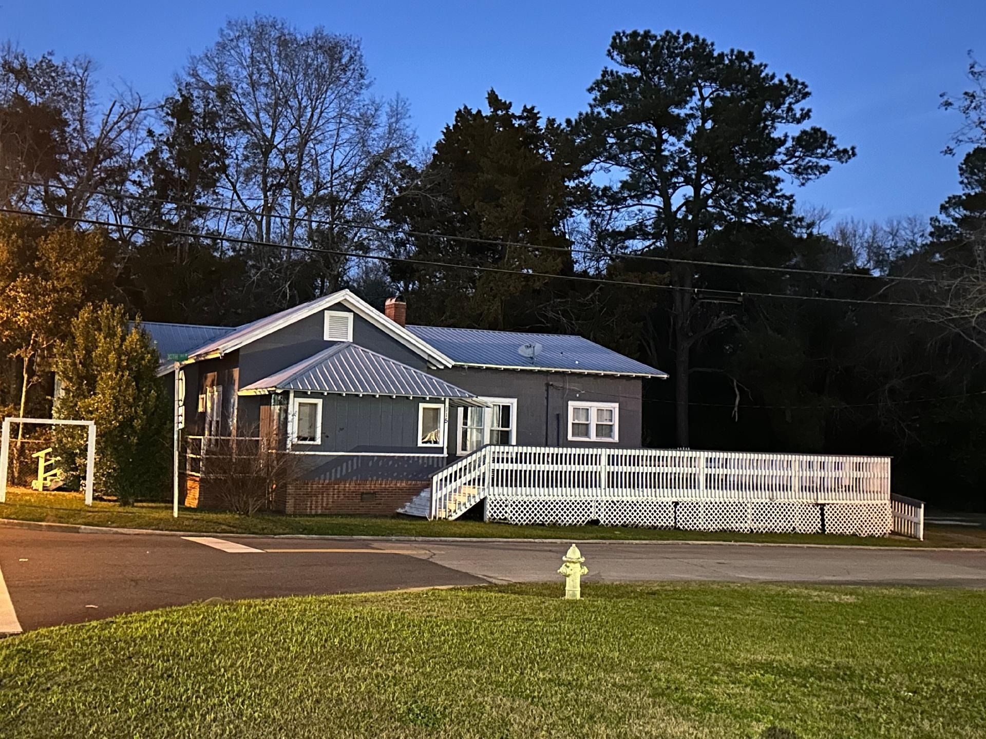 Blue house at dusk with lit porch, white fence, and a grassy foreground near the road.