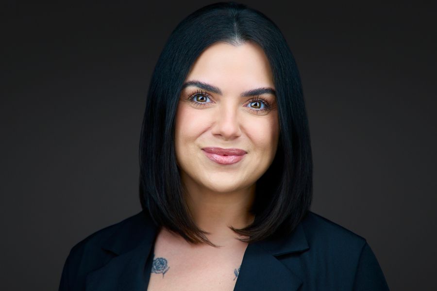 Woman with short brown hair, smiling, wearing a dark blue silk blouse, and a small necklace; dark background.