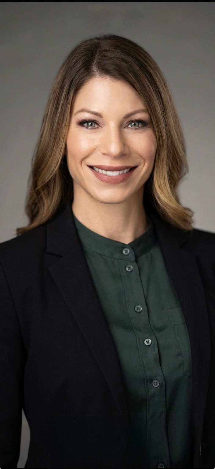Woman with long dark hair smiles at the camera, wearing a grey blazer over a black shirt, against a neutral background.
