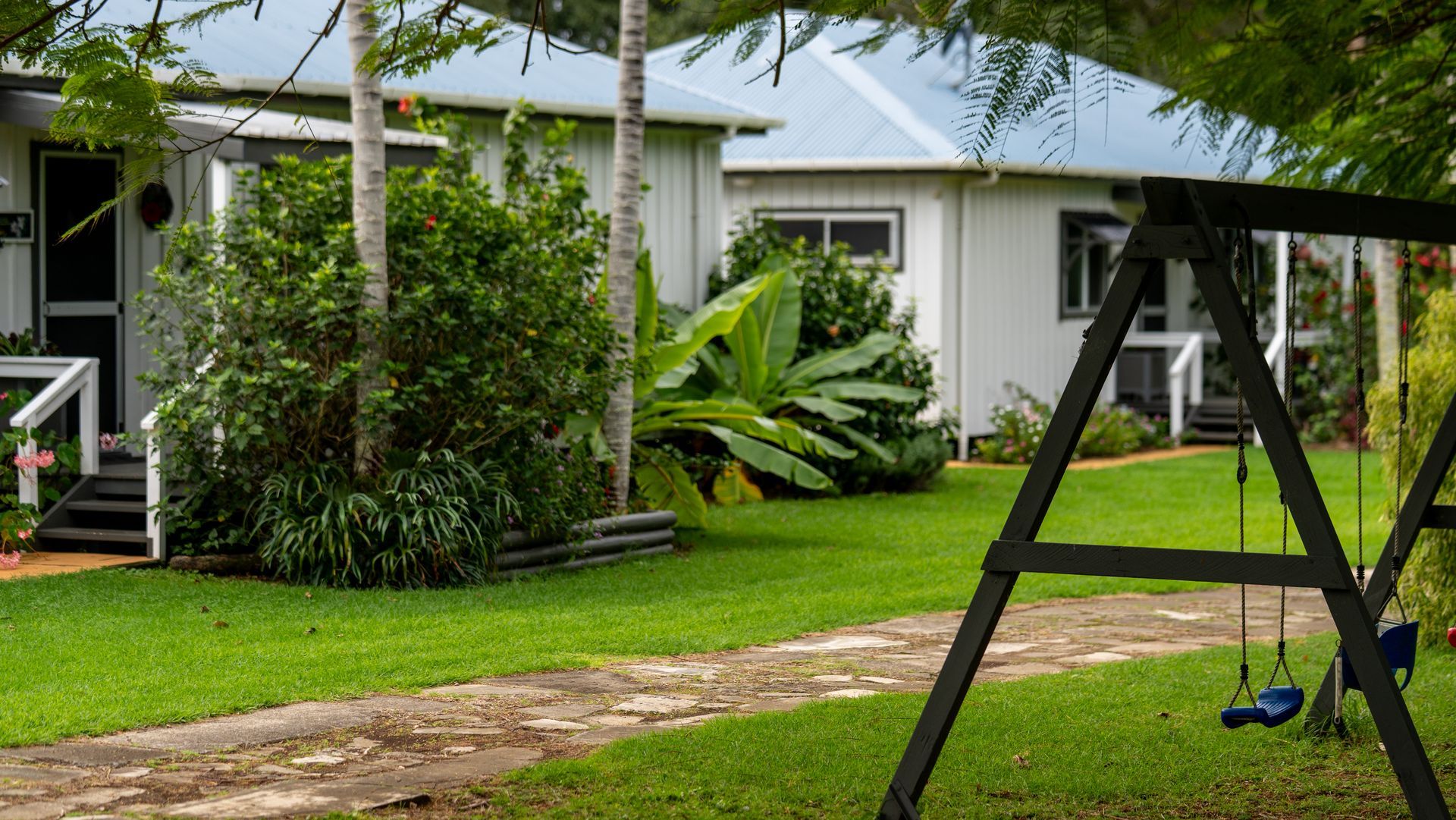 White house with green lawn, palm trees, and a black swing set in the foreground.