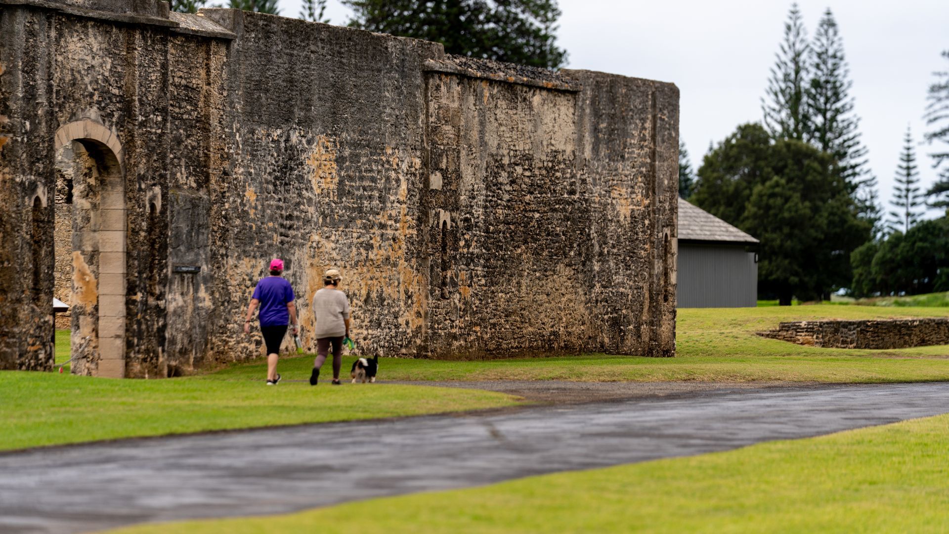 Two people walking by a moss-covered stone wall on a rainy path in a park