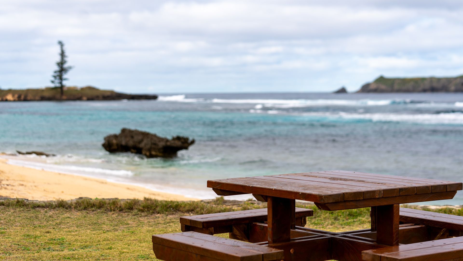 Wooden picnic table by a sandy beach with turquoise water and a rocky shoreline at low tide