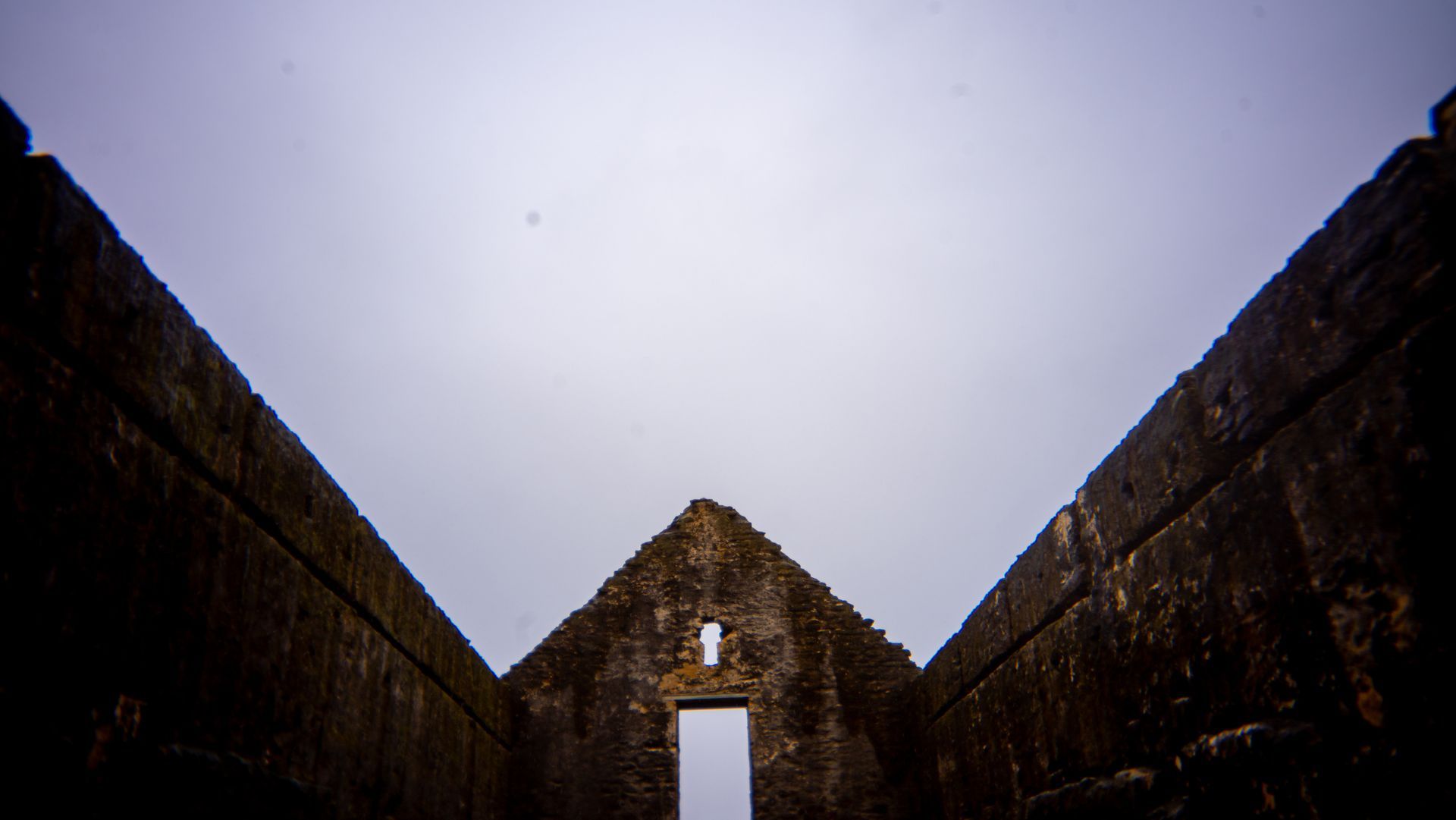 Ruined stone structure with a triangular roof opening, seen from a dark corridor under a cloudy sky.