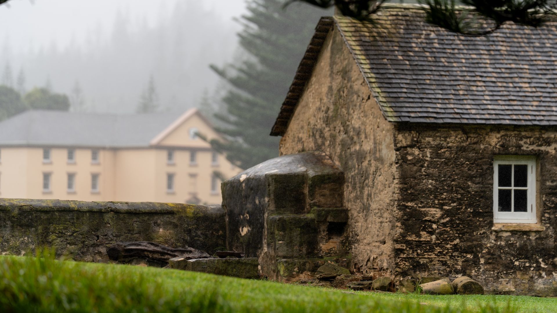 Old stone cottage in a misty rural setting with a beige house and evergreen trees in the background