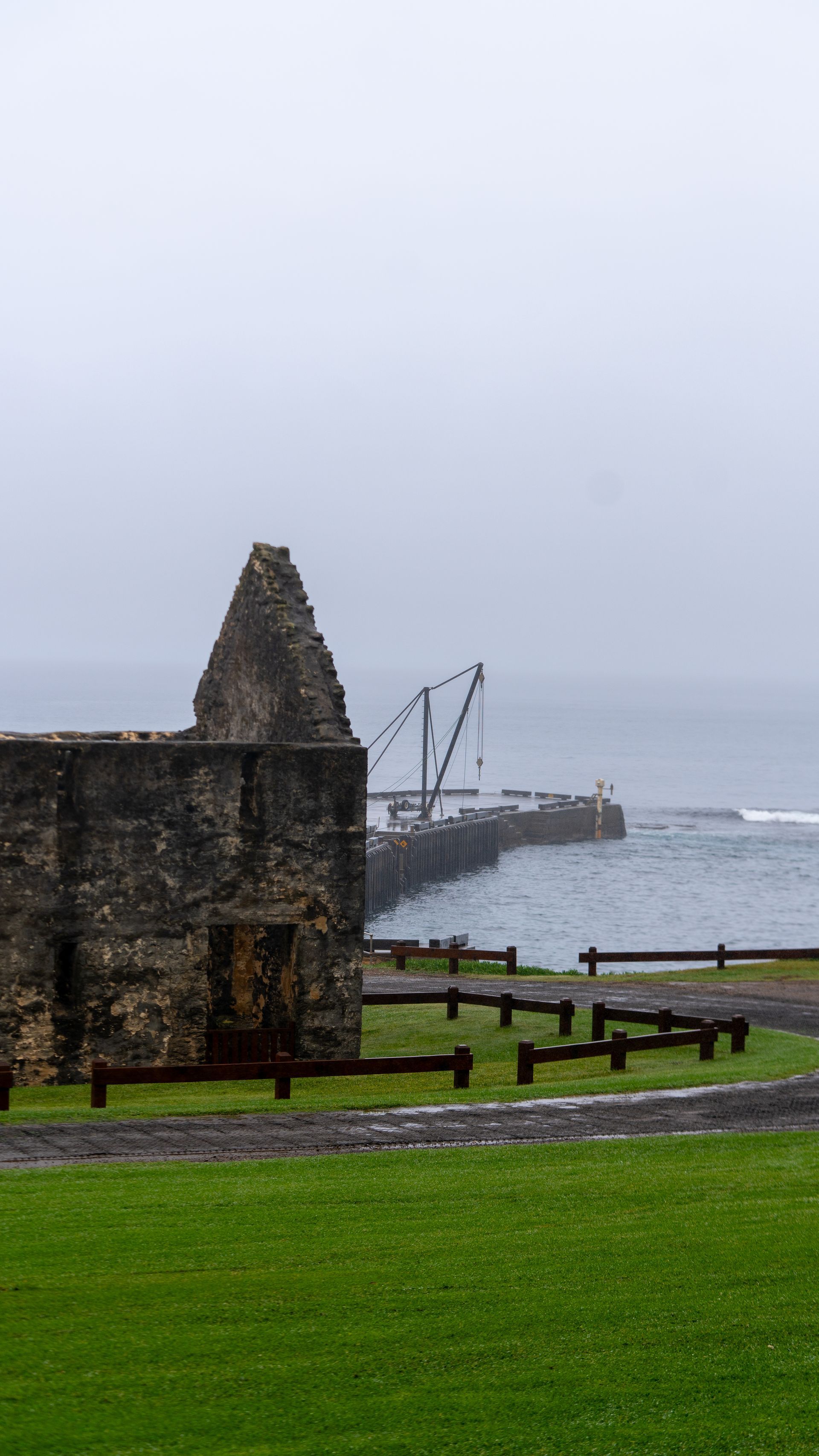 Stone ruins by a misty seaside harbor with a green grassy foreground and overcast sky