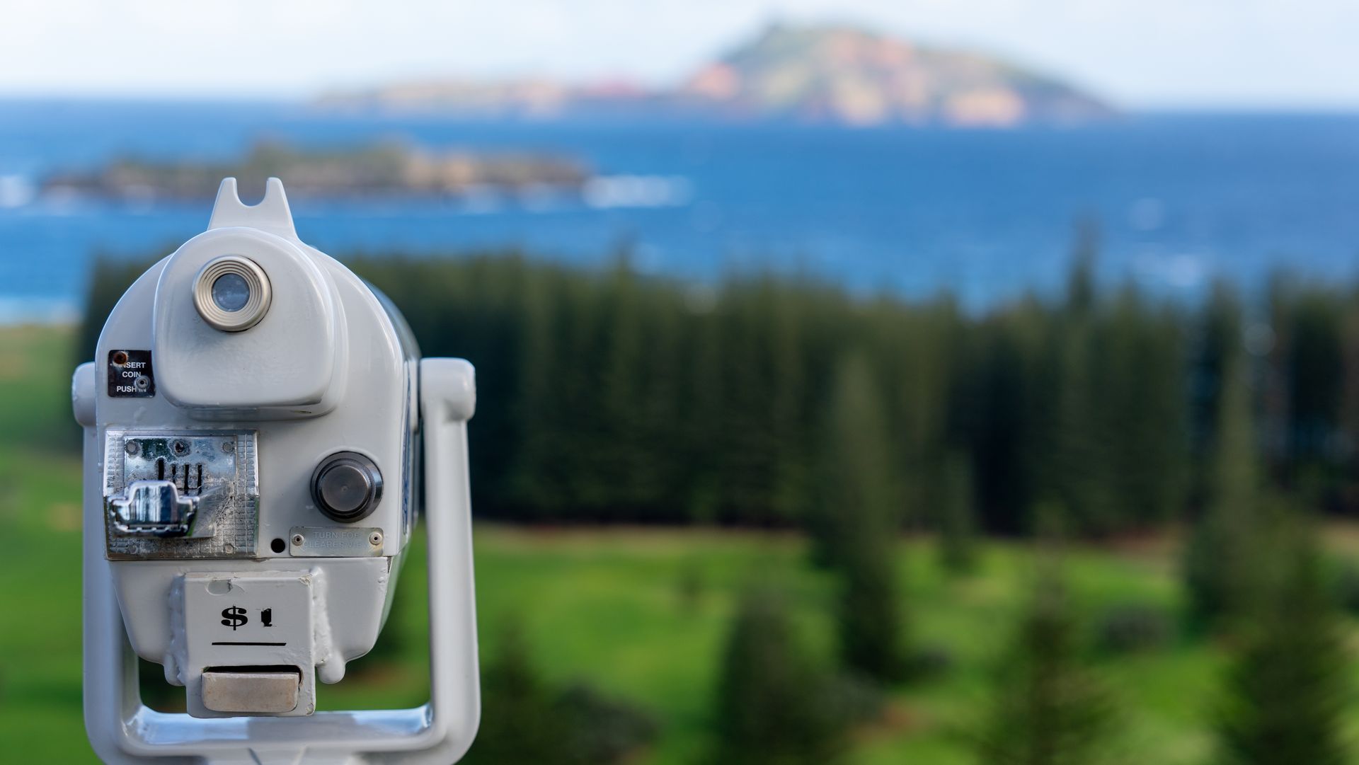 White coin-operated viewer overlooking a blue sea and distant island on a blurred, green coastline