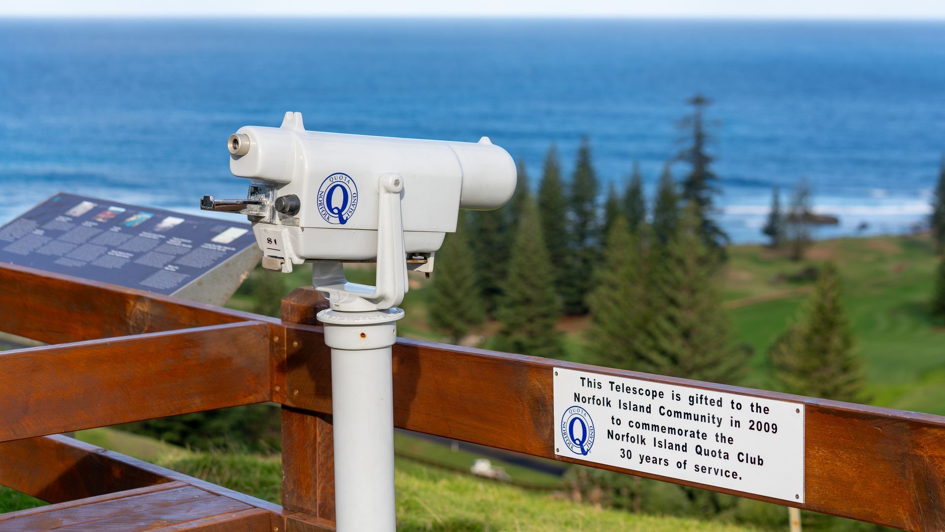 Coin-operated binoculars beside a seaside lookout, with ocean and trees in the background.