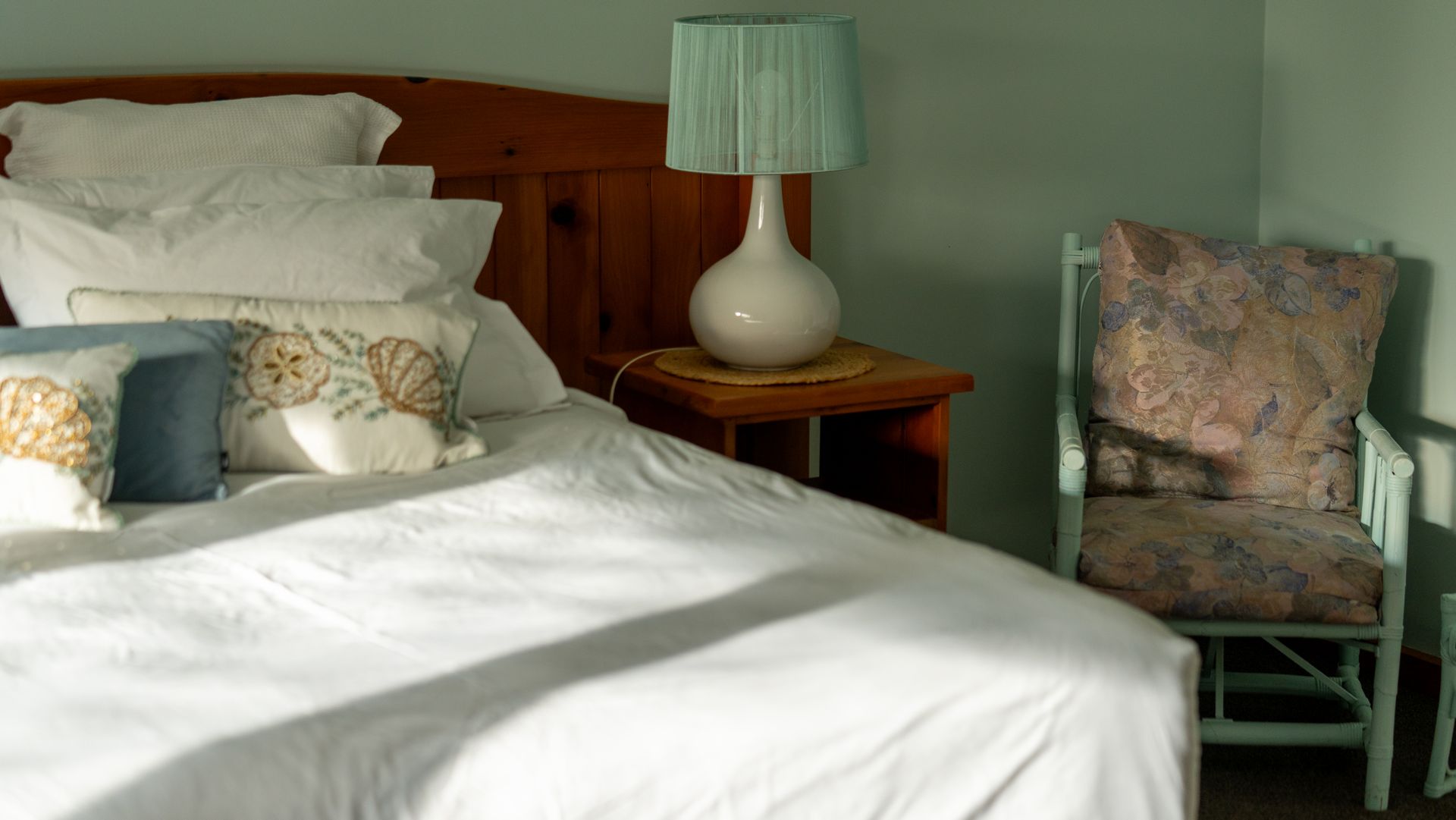 Bed with white linens, pillows, bedside lamp, and upholstered chair in a softly lit room.