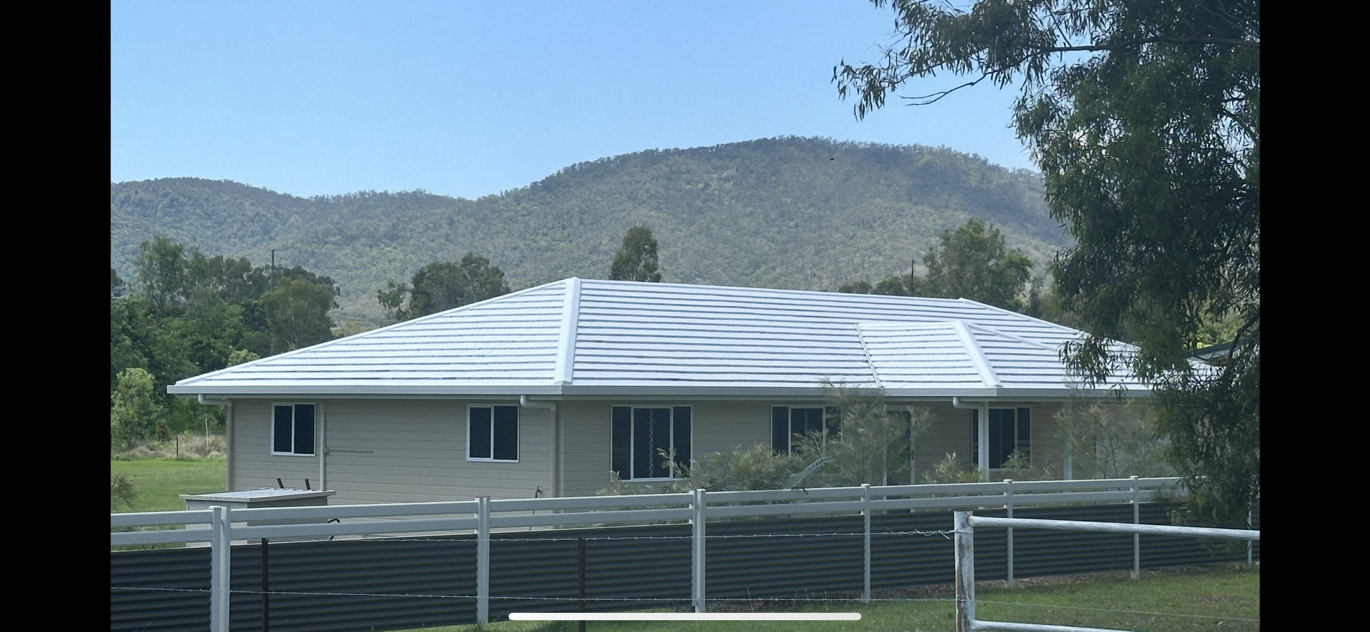 A house with a wavy white roof and windows, a fence in front, and mountains in the background — Roof Tek CQ in Gracemere, QLD