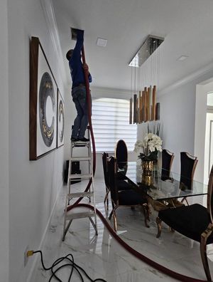 Person on ladder cleaning air vent in a dining room. Red hose connected, white walls, marble floor, chandelier.