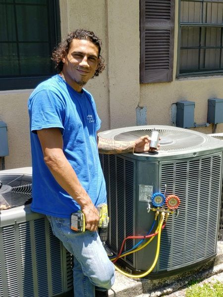 Man in blue shirt, working on an AC unit outdoors. He holds a tool and gauge.