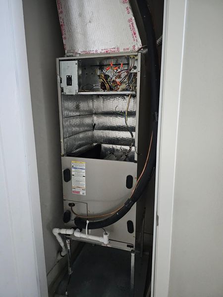 HVAC unit in a closet. Metallic and insulated surfaces, various wires and pipes. White walls surround.