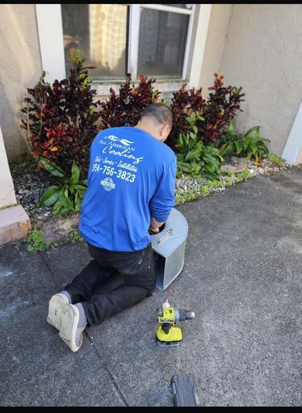 Man in blue shirt, kneeling, working with metal object on concrete near bushes and a window.