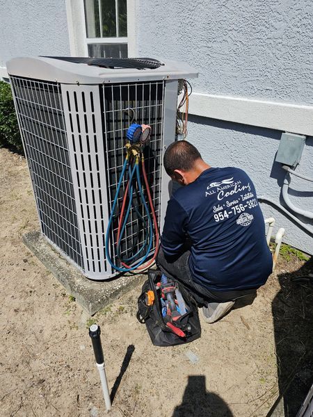 HVAC technician kneels, connecting gauges to an air conditioning unit outside a light blue building.