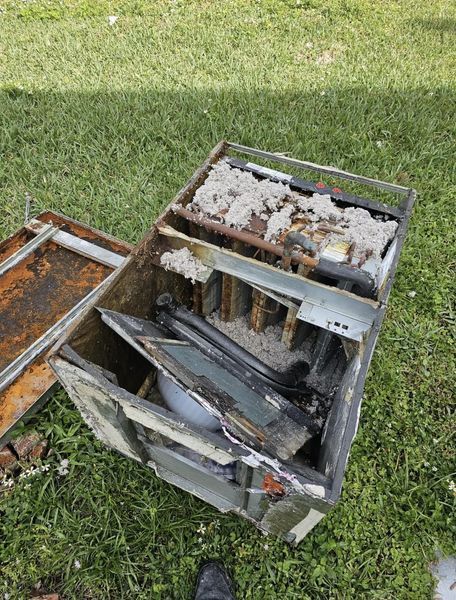 Damaged, open metal box on grass, with burnt components and insulation visible. Rust and debris present.
