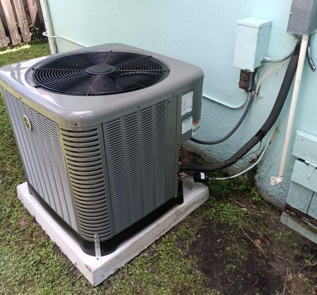 Air conditioning unit, gray, sitting on a concrete pad, next to a pale blue building.